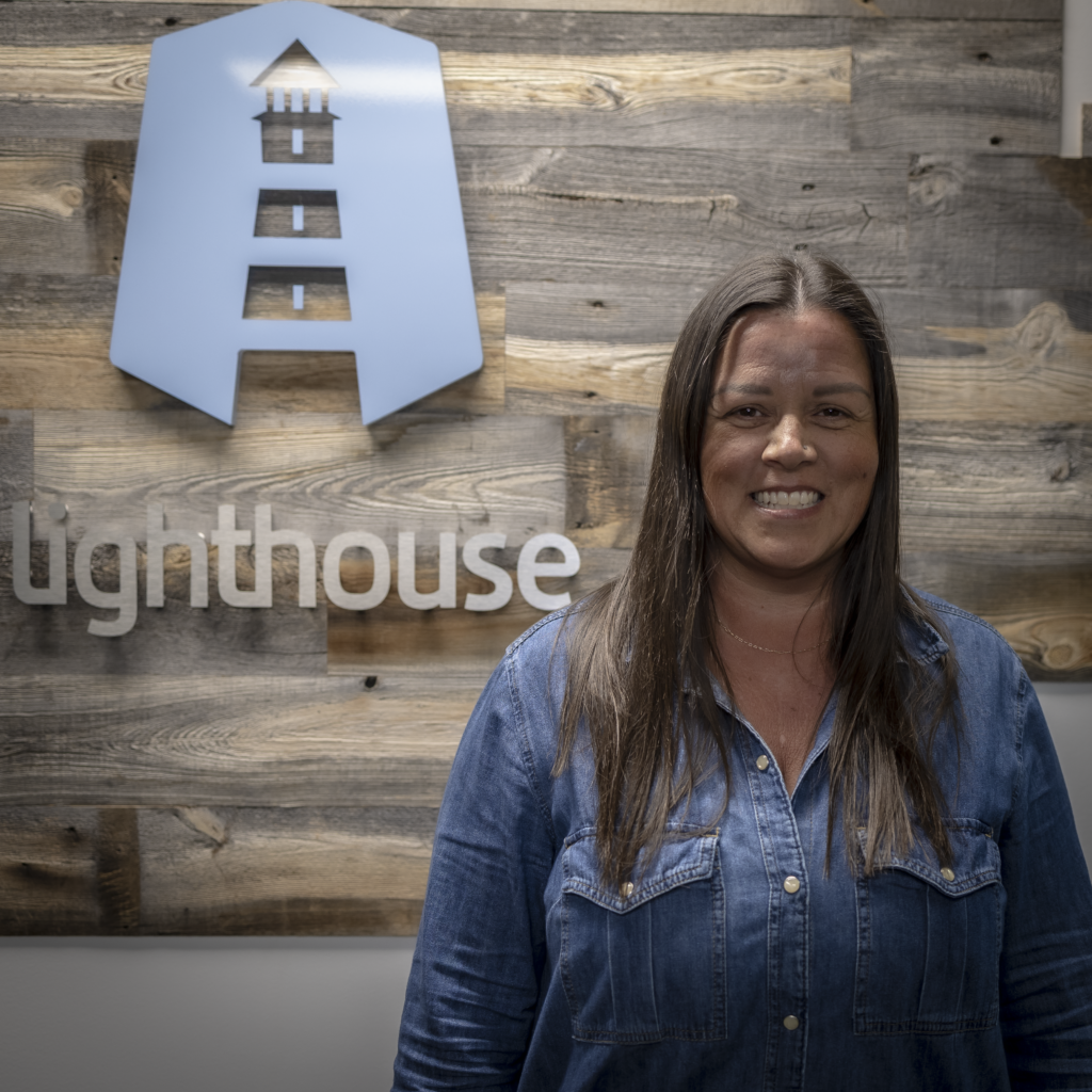 Office Manager, Tammy Stang smiling in front of the Lighthouse sign on wooden wall